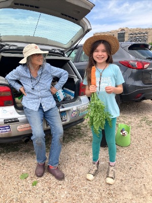 Ila leaning back on her car looking at a smiling young girl holding a carrot